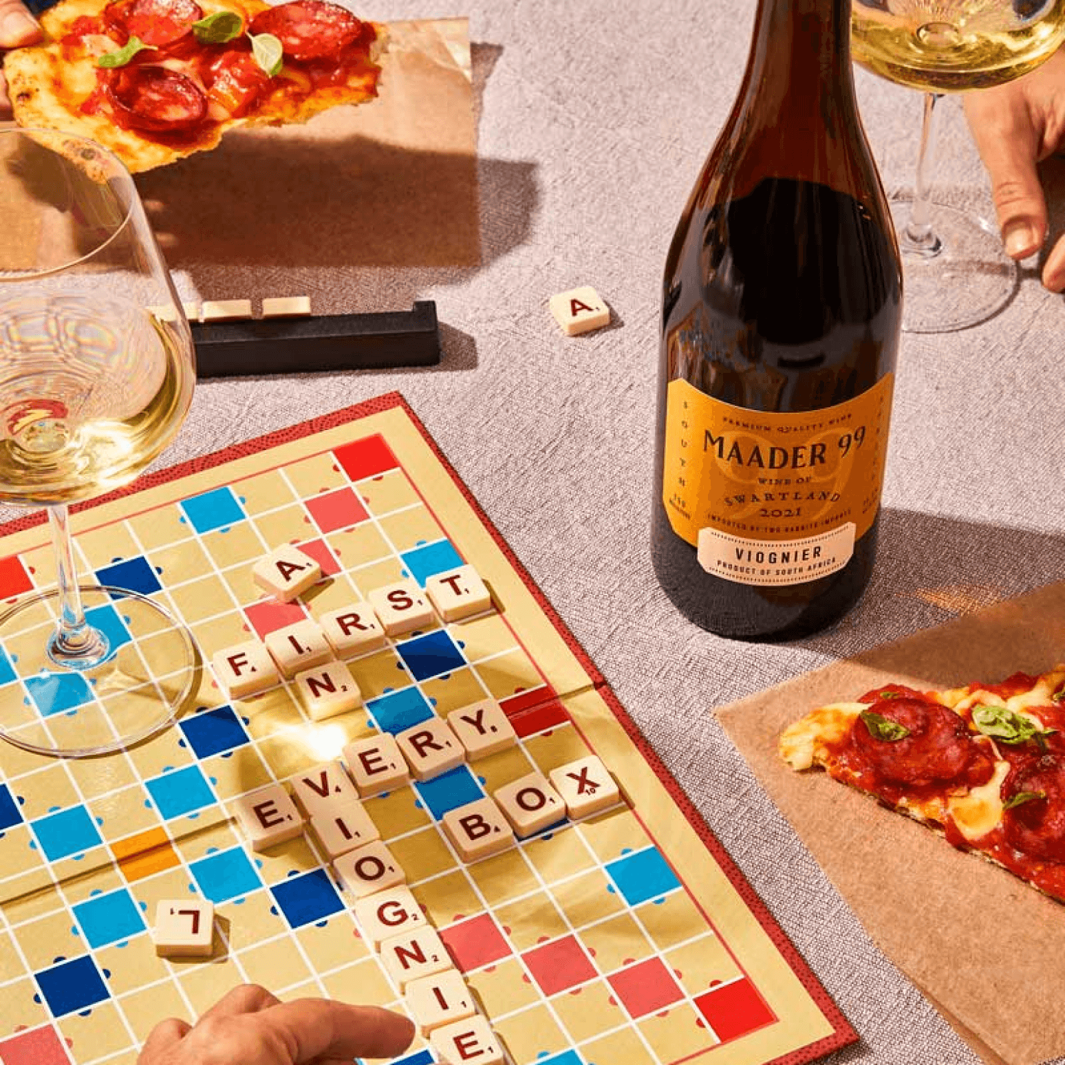 Graphic, a bottle of Maader 99 Viognier sitting on a table with pizza, wine glasses, and a Scrabble board. The pieces on the Scrabble board spell out a first in every box Viognier.