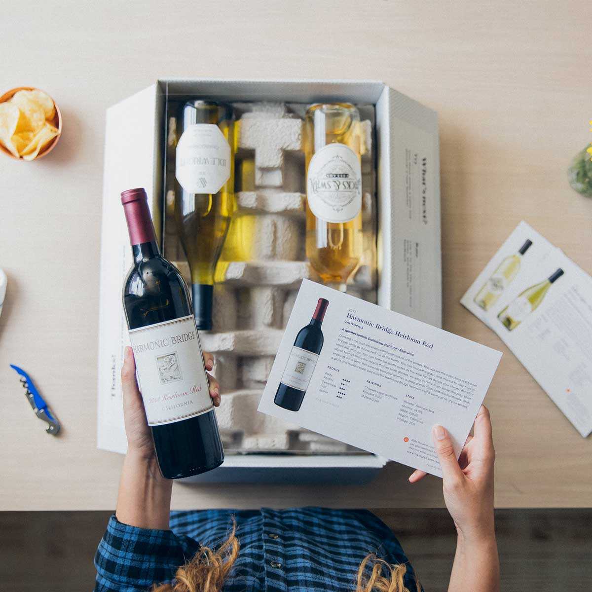 Graphic, a Firstleaf box of wine and descriptive wine cards on a table seen from above with a woman's hands holding a bottle of red wine and it's wine card.