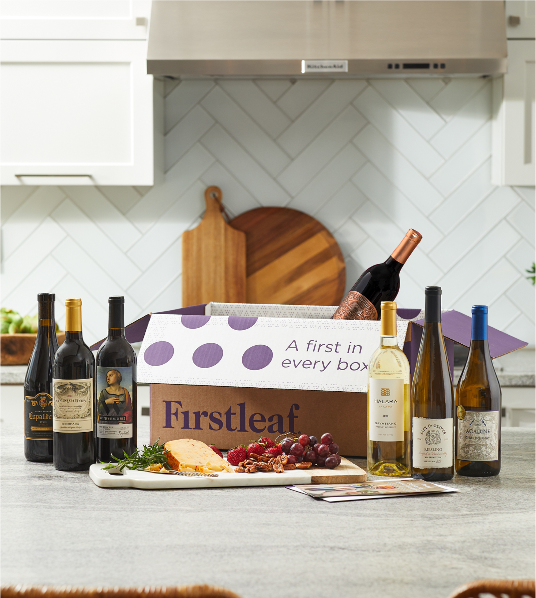 Image, an open Firstleaf box, six bottles of wine, and a fruit and cheese platter on a kitchen counter.