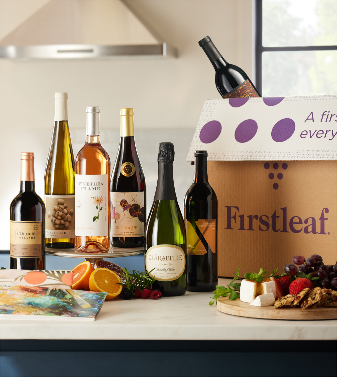 Image of a countertop with 6 wine bottles lined up next to a Firstleaf delivery box, with a 7th bottle peeking out of the box and a fruit and cheese plate and oranges.