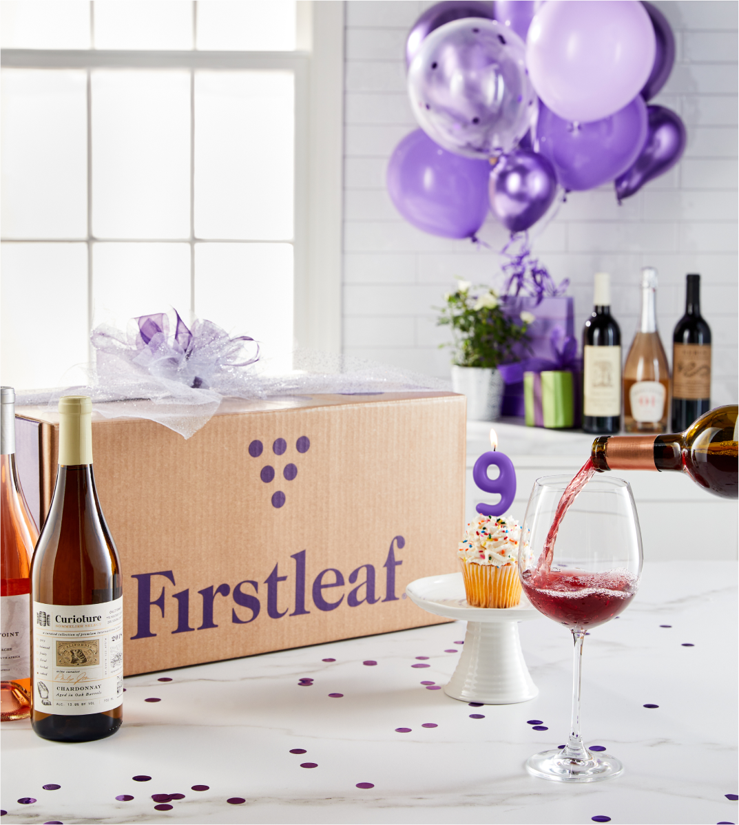 Image, a Firstleaf box with a purple bow on a kitchen counter, bottles of wine, a cupcake with a candle shaped like the number nine, and a glass of red wine being poured.